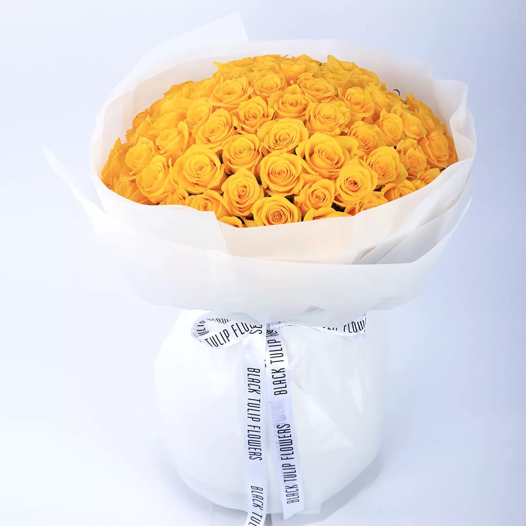 100 stems of vibrant yellow roses arranged in a white wrap, tied with a white ribbon.