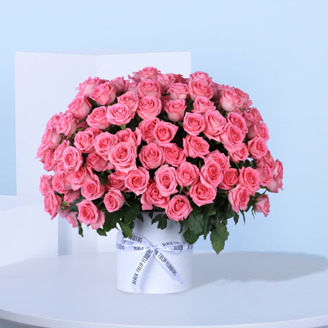 A white circular box filled with light pink roses, arranged in a circular pattern tied with white ribbon.