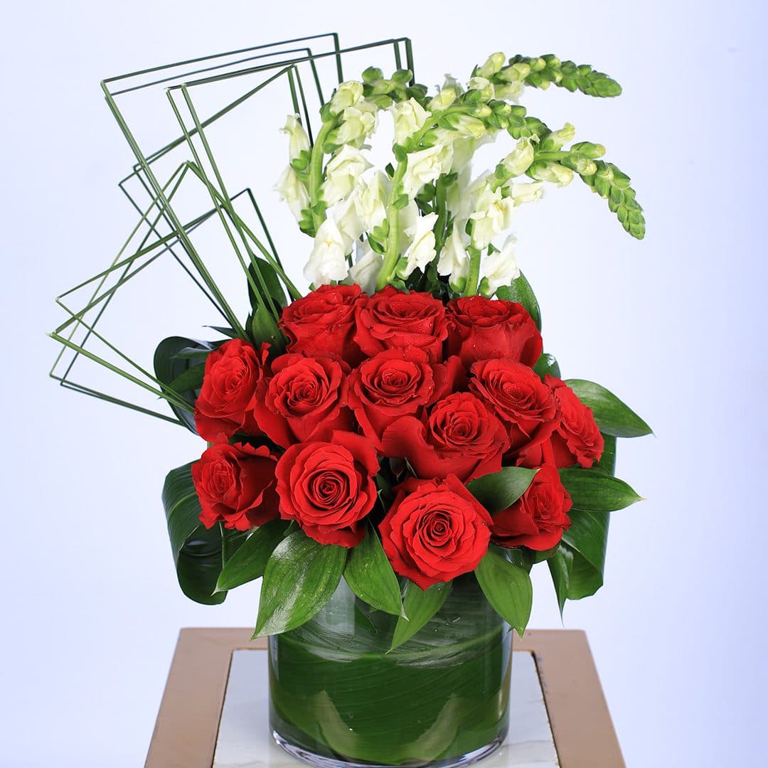 A floral arrangement in a glass vase, with red roses, white snapdragons, greenery, and geometric wire accents.