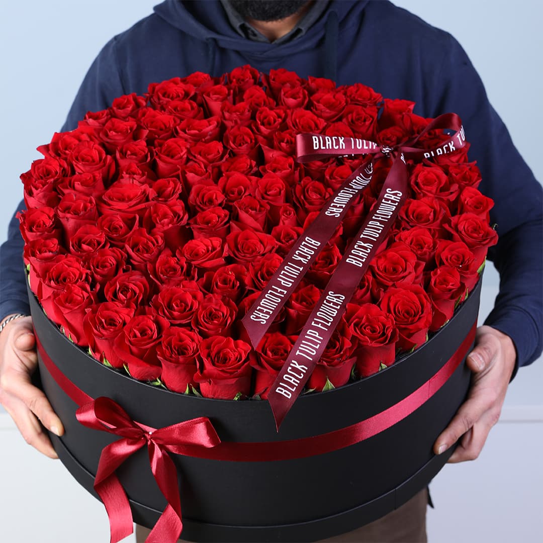 Luxury Valentine flower box featuring 100 red roses arranged in a black round box with red ribbon bow.