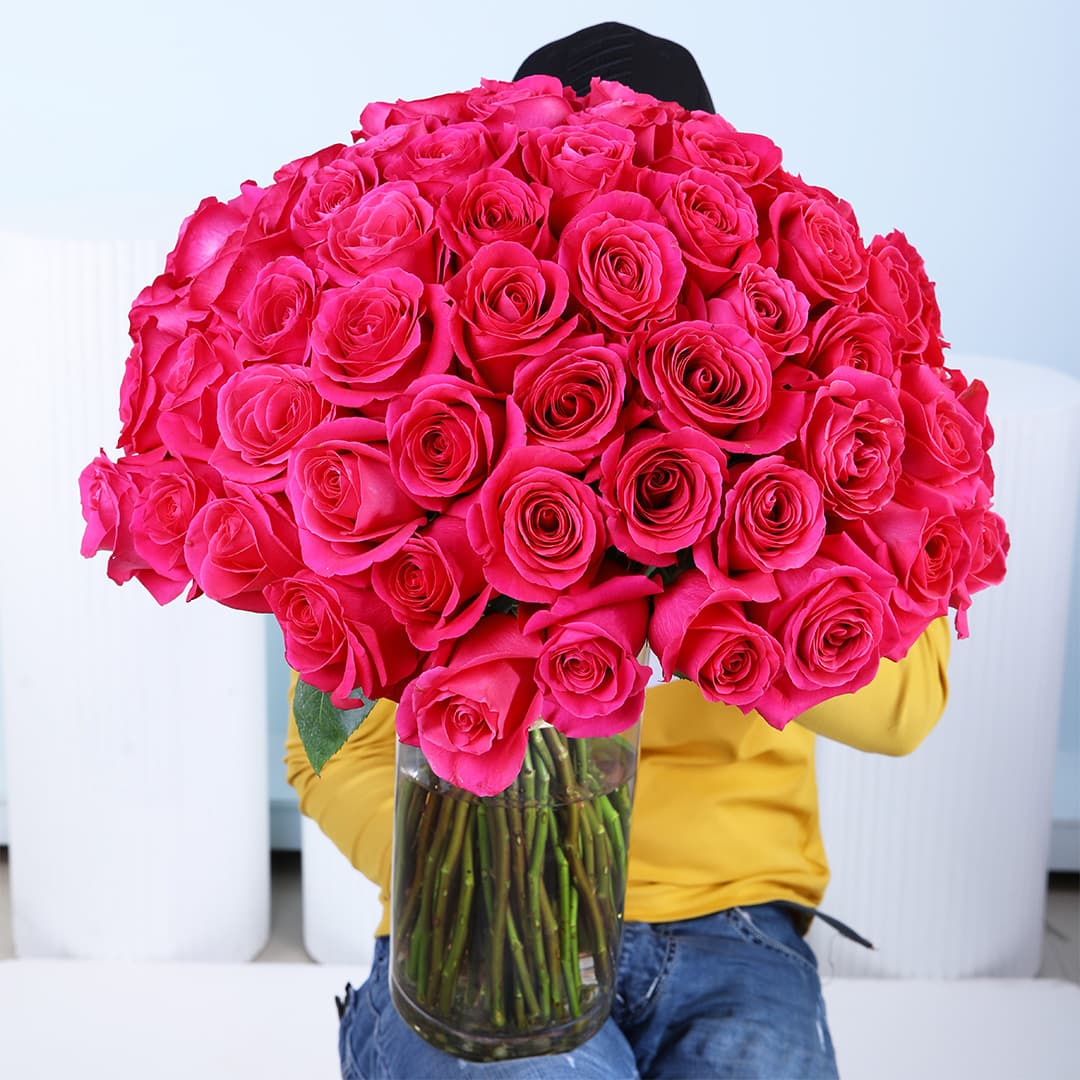 A large, full bouquet of 100 purple roses with visible green stems, meticulously arranged in a tall, clear glass cylindrical vase and tied with a natural-colored fabric.
