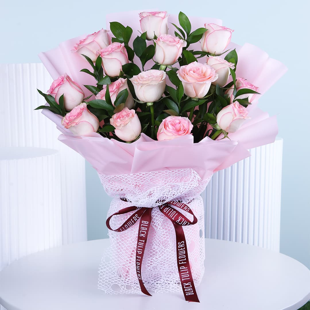 A beautiful bouquet featuring soft pink roses, hand-tied and wrapped in light pink, displayed on a white table.