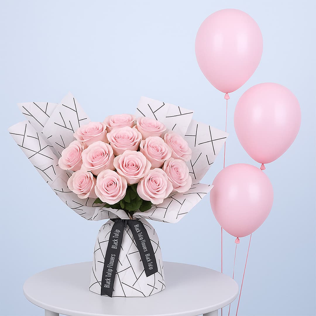 A bouquet of light pink roses wrapped in white paper with a black geometric pattern along with pink balloons.