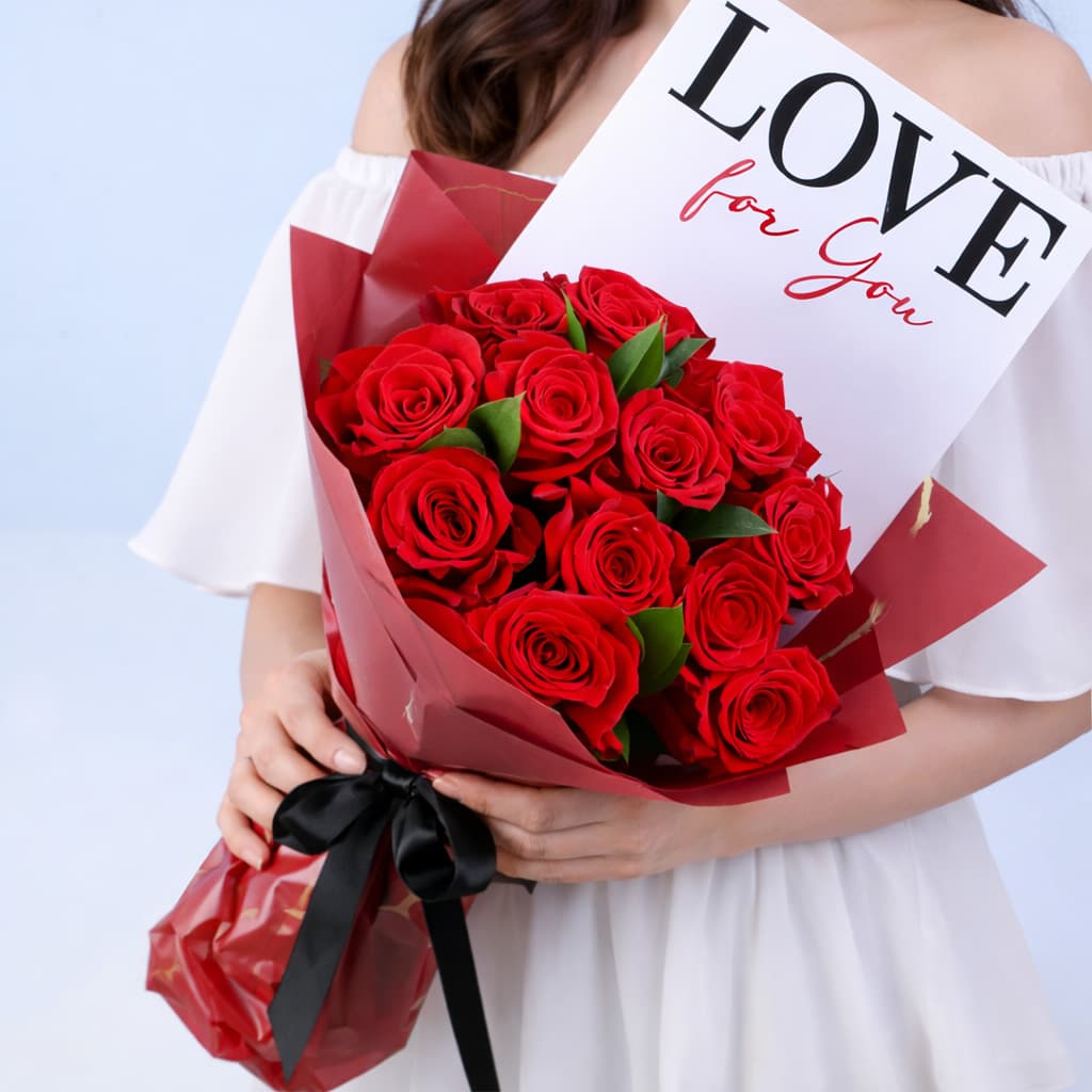Bouquet featuring red roses with green leaf accents in premium red wrapping, finished with a LOVE message card.