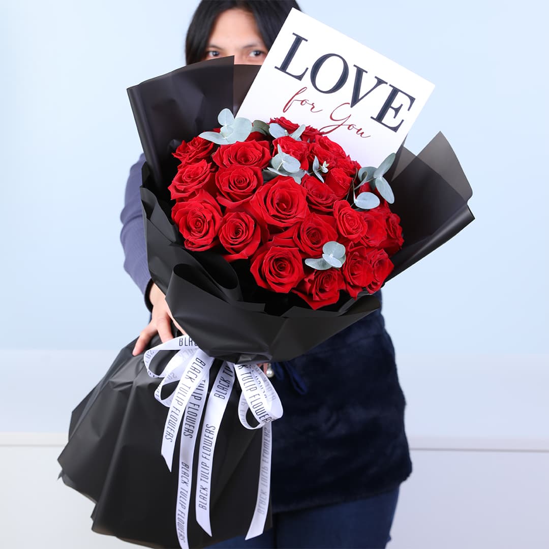 A bouquet of red roses and eucalyptus in black wrapping, finished with a Love For You card.