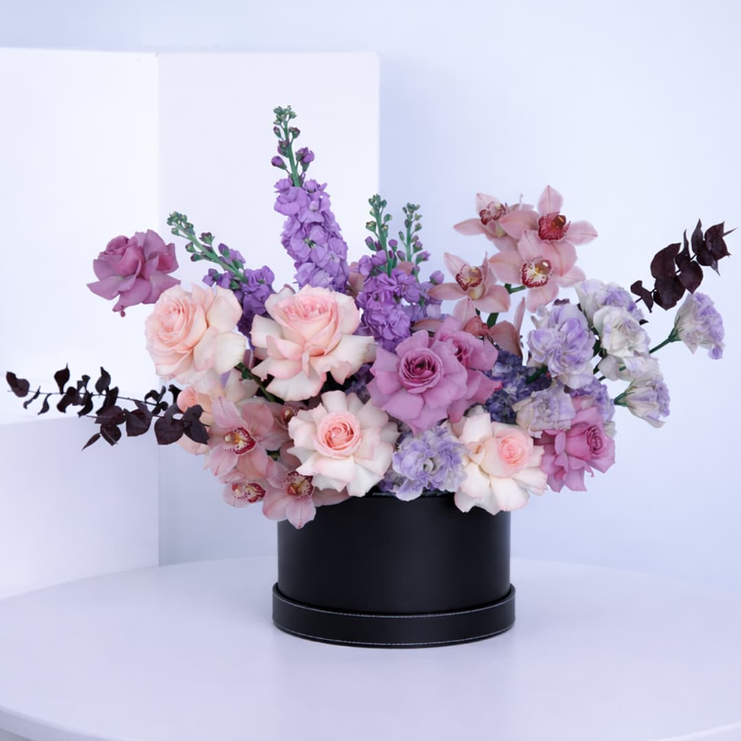 A luxurious mix of pink roses, purple roses, cymbidium, purple matthiola, purple lisianthus, blue hydrangeas, and eucalyptus, elegantly arranged in a sleek black box.