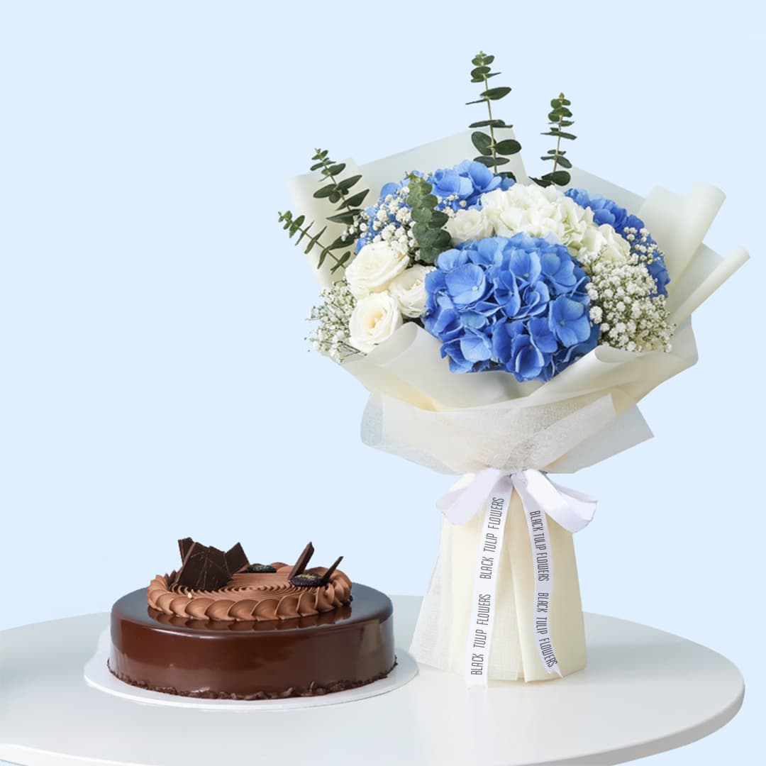 A bouquet featuring blue hydrangeas, white roses, baby's breath, and eucalyptus, wrapped in white and tied with white ribbon. A rich chocolate cake with a glossy glaze and chocolate frosting is adorned with chocolate bits.