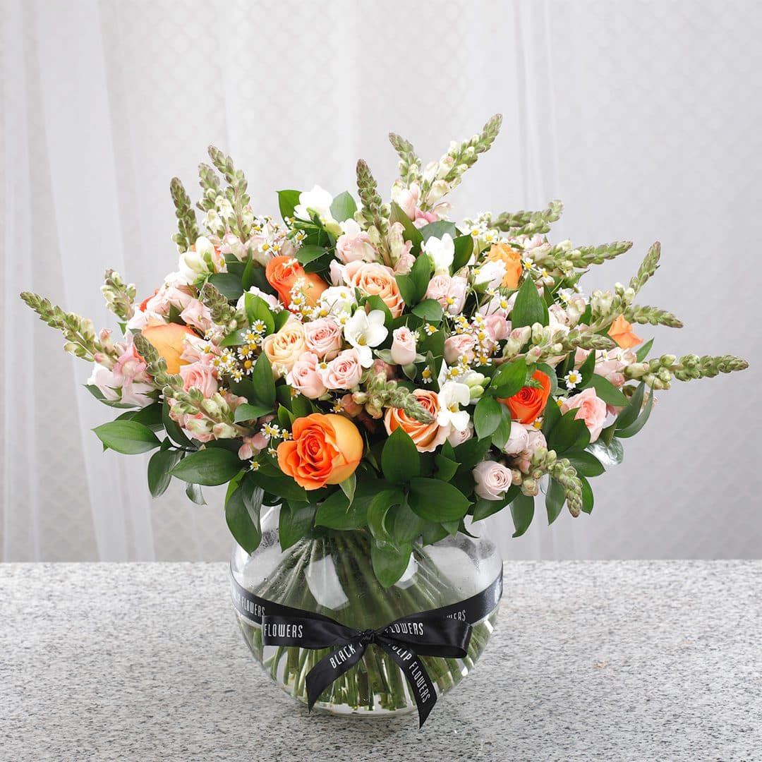 A clear glass vase featuring peach and orange roses, white snapdragons, and delicate blooms, tied with a black ribbon.