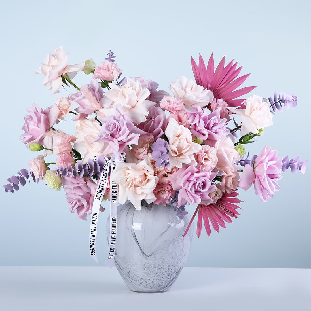 Light pink and purple roses with lisianthus and premium fillers in a marble-effect glass vase arrangement, finished with a white ribbon.