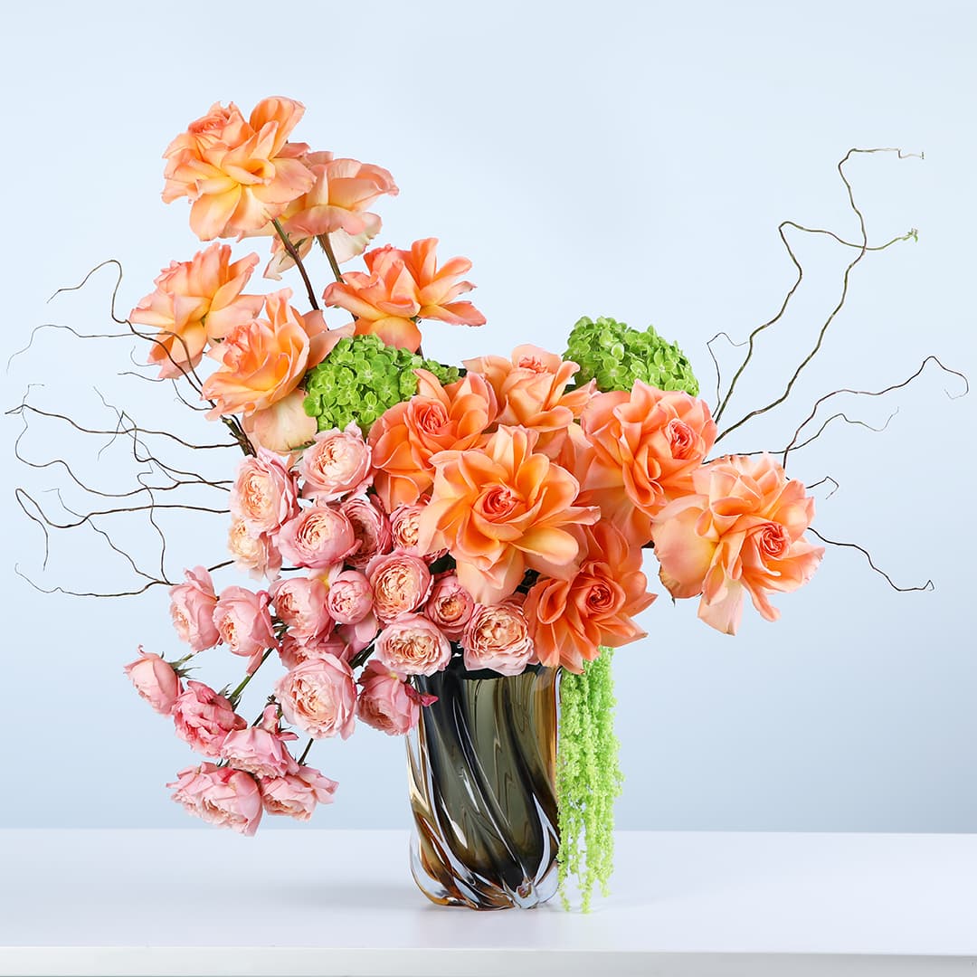 Floral vase featuring roses, baby roses, green hydrangeas, and cascading amaranthus in a designer vase with artistic branches.