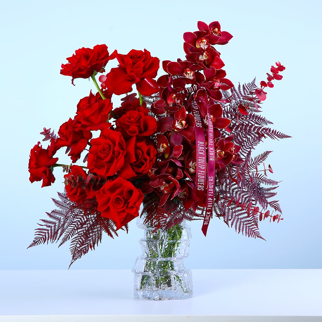 A glass vase featuring red roses, red cymbidium orchids, and decorative foliage, styled for luxury decor and gifting.