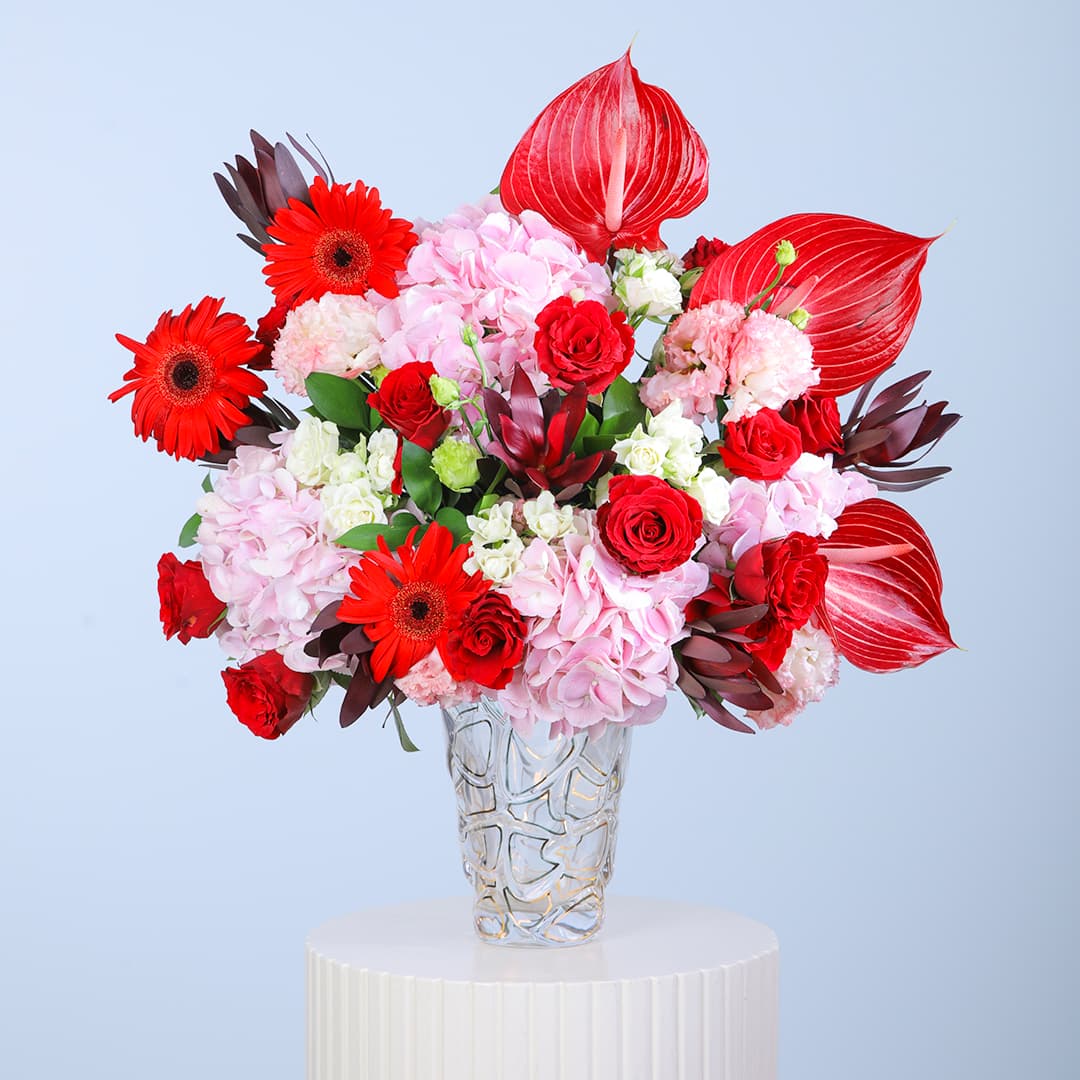 A floral arrangement in glass vase featuring red gerberas, pink hydrangeas, red roses, and striking anthurium leaves.