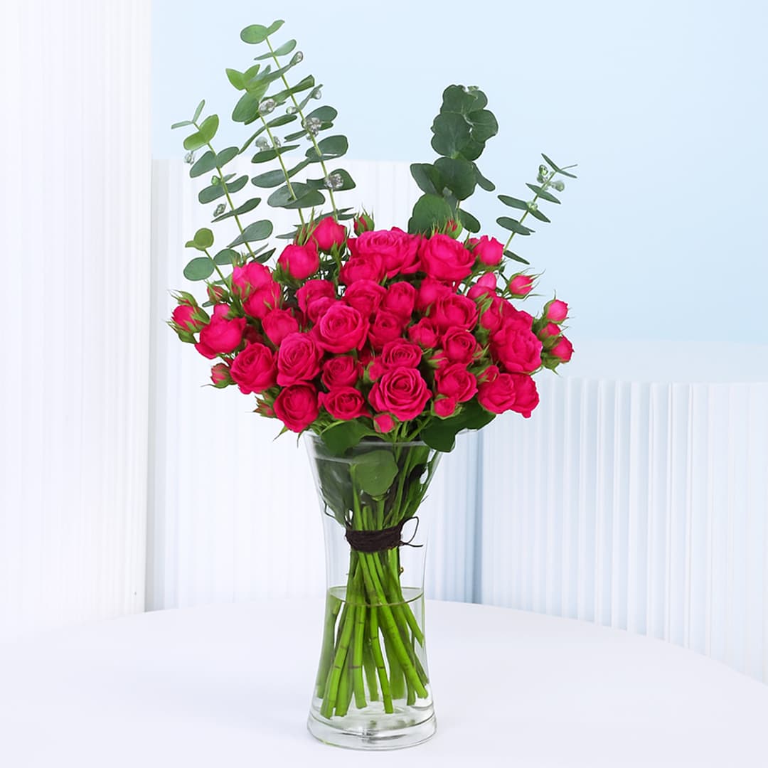 A bunch of 20 vibrant dark pink spray roses in a clear glass vase, with tall green eucalyptus leaves providing complementary foliage.