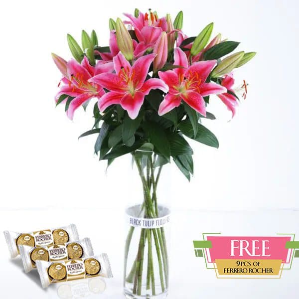 A bunch of pink lilies in a glass vase, accompanied by a box of Ferrero Rocher chocolates.