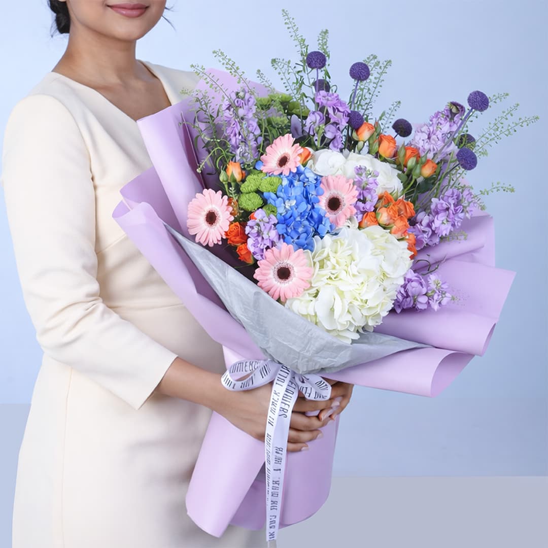 A vibrant mixed floral arrangement featuring pink Gerbera, white and blue Hydrangea, purple Mattiolla, Caspedia, green Chrysanthemums, and orange Baby Roses in a sleek gift box.