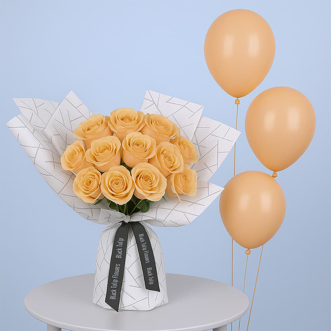 A bouquet of peach roses arranged in a white wrap with a black geometric design. It is tied with a black ribbon accompanied with peach-colored balloons.