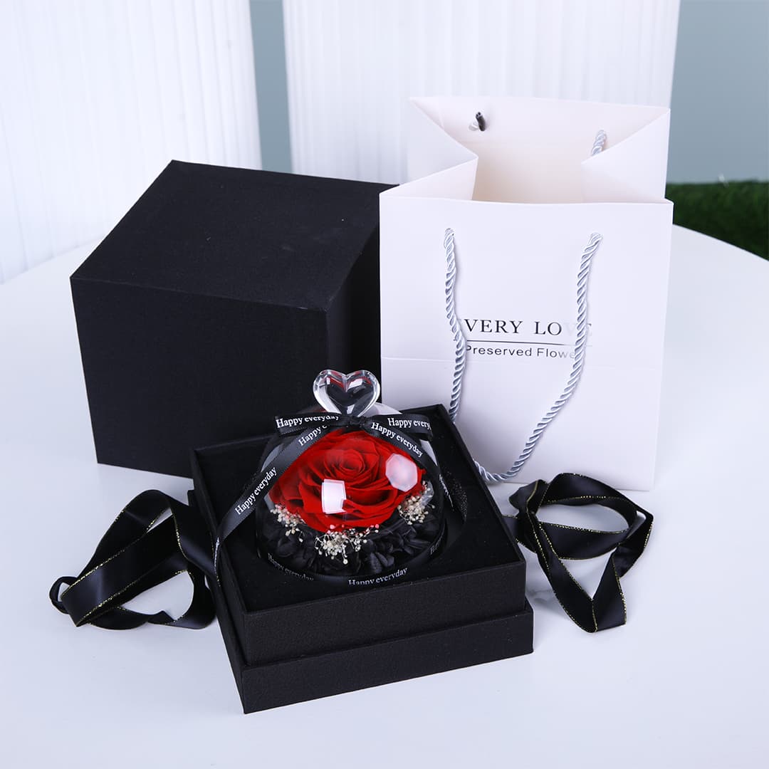 A preserved red rose in a glass dome inside a black gift box, with a white gift bag and black ribbon