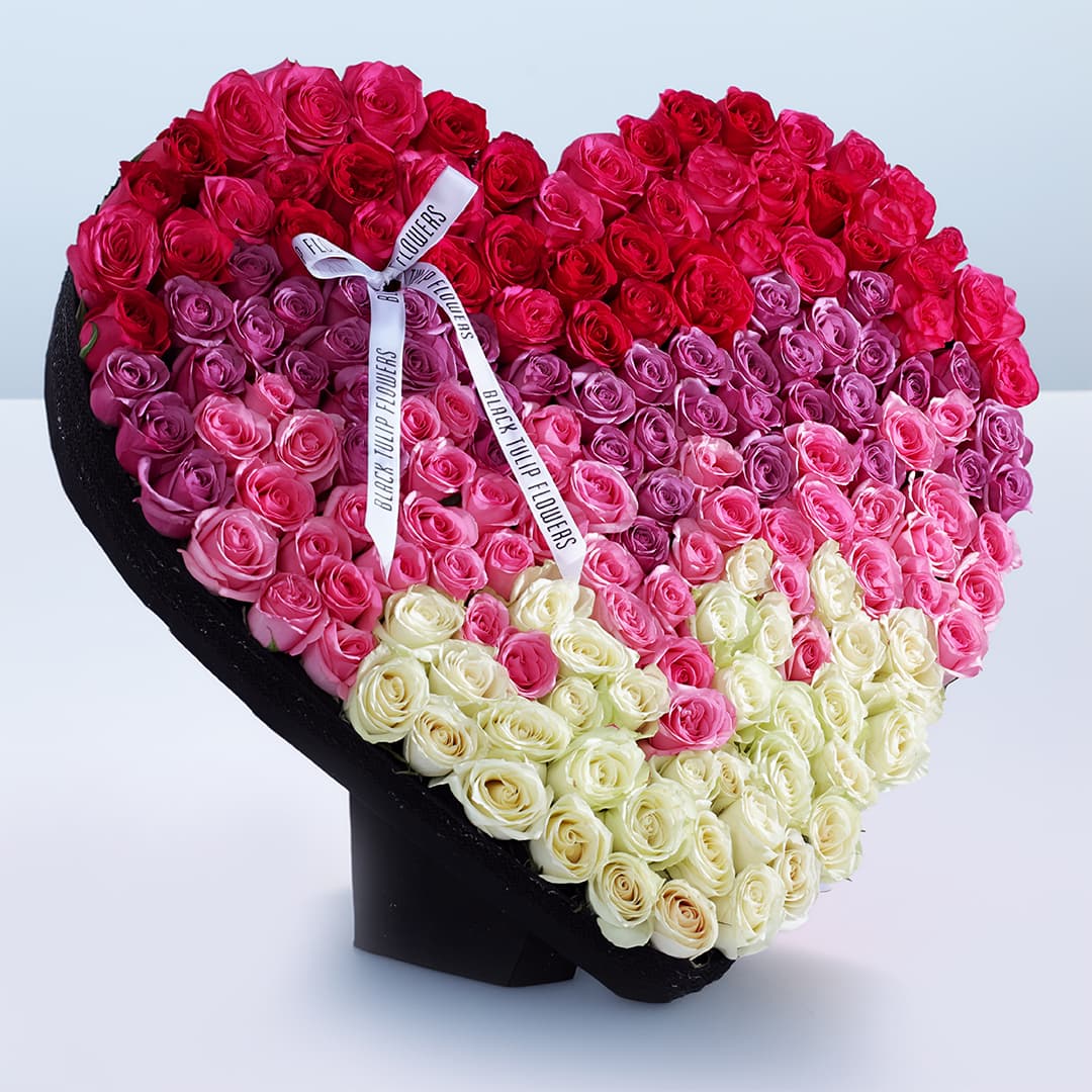 180-200 mix coloured roses arranged in a heart shape, adorned with a white ribbon.