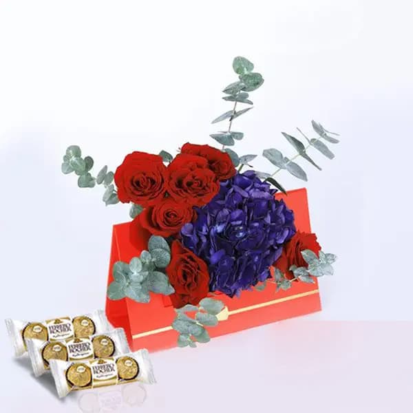 A red card with red roses, blue hydrangeas, and eucalyptus leaves, accompanied by a box of chocolates on the table below.