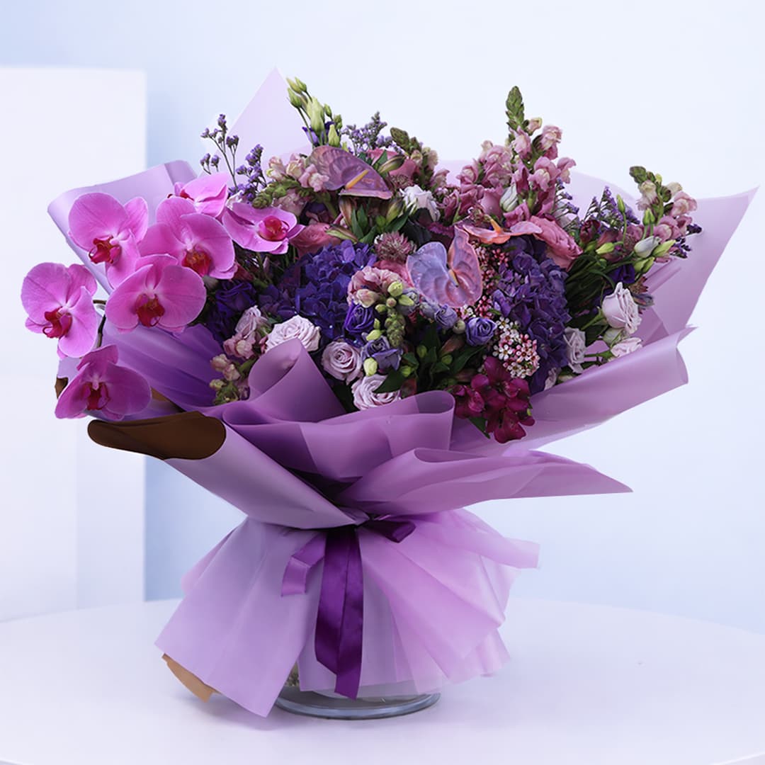 Bouquet of purple and pink flowers, including orchids, roses, and hydrangeas, wrapped in purple paper.