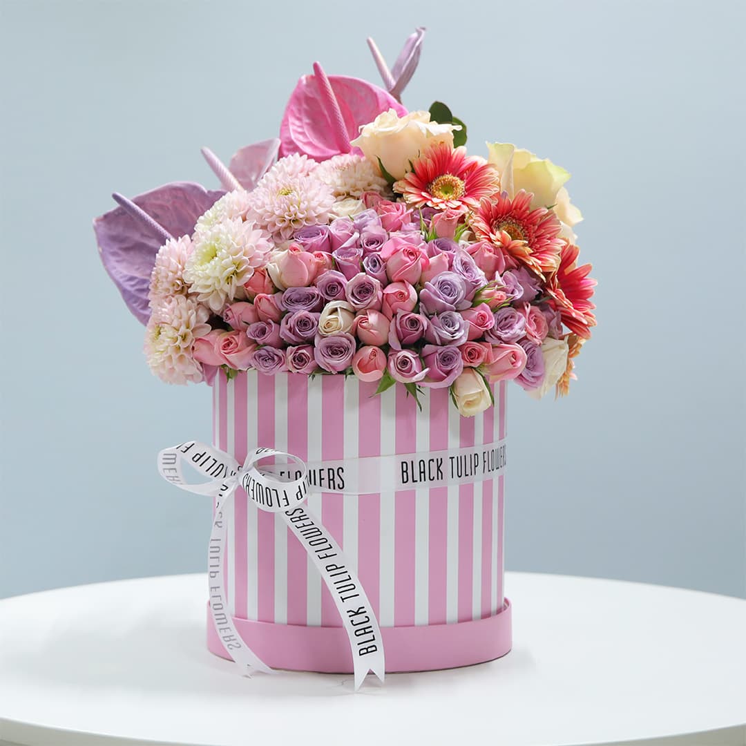 A pink striped cylindrical box filled with pink roses, purple spray roses, white chrysanthemums, pink anthuriums, and orange gerberas.