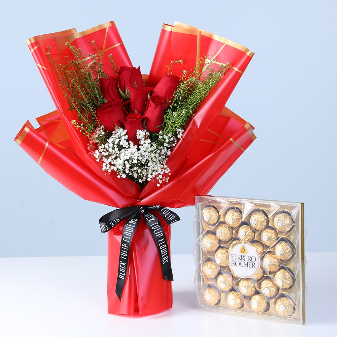 Bouquet of red roses with white filler flowers and green foliage in pink wrapping, with a Ferrero Rocher chocolate box.
