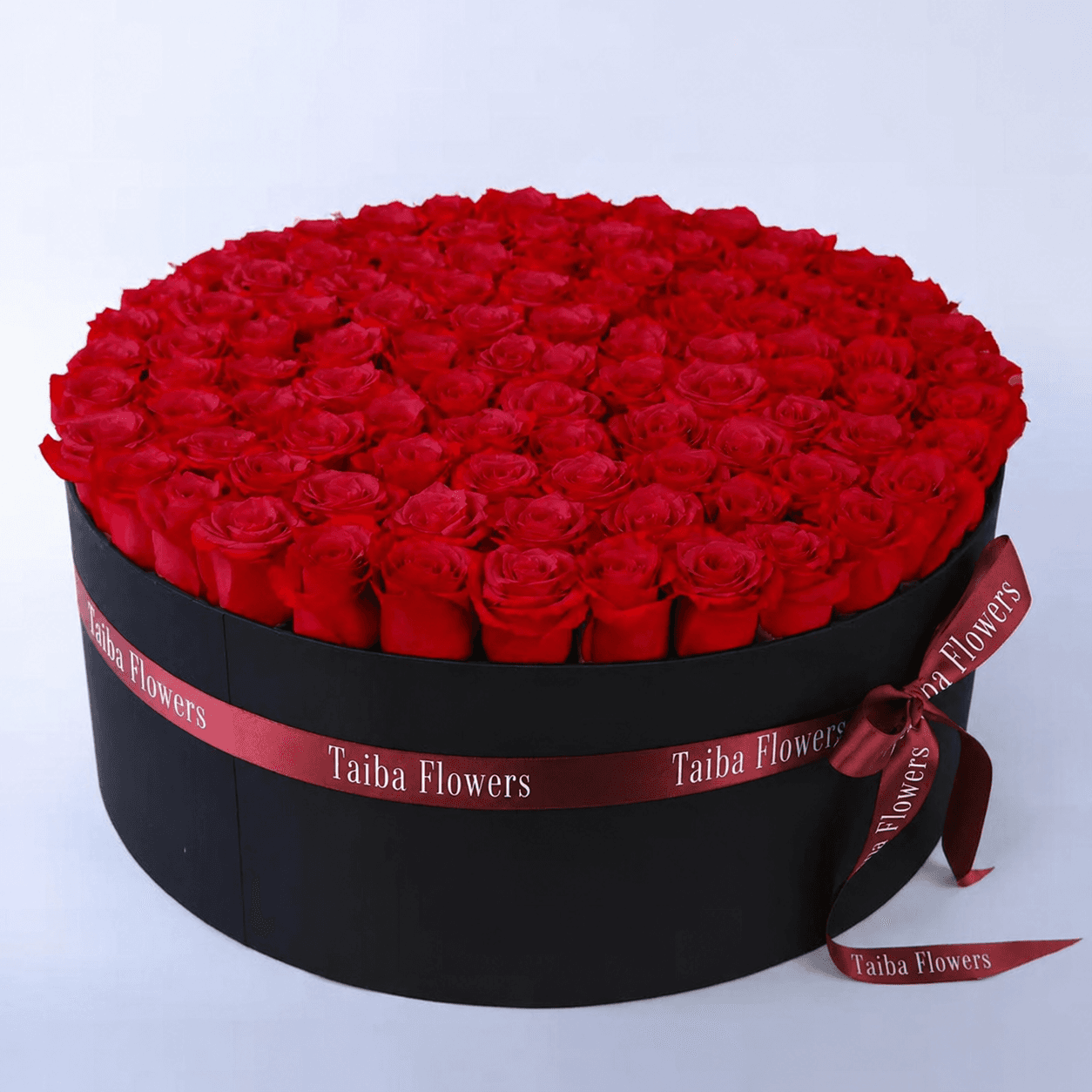 A sleek black circular box adorned with a red ribbon, filled with a stunning arrangement of vibrant red roses.