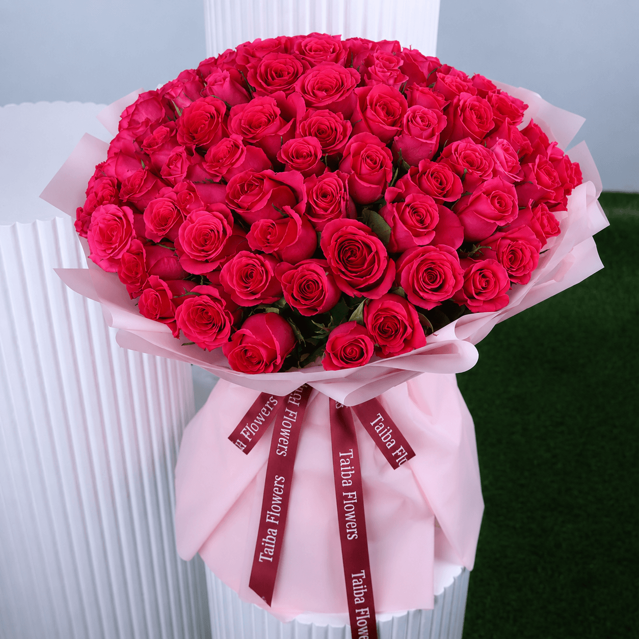 A rounded bouquet of Dark pink roses wrapped in pink paper with a "BLACK TULIP FLOWERS" ribbon, against a white and green background