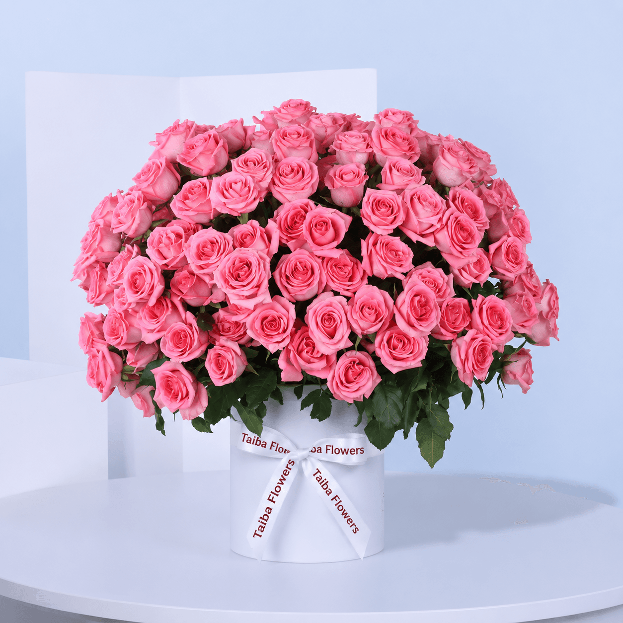 A white circular box filled with light pink roses, arranged in a circular pattern tied with white ribbon.